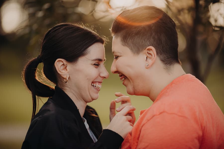 The couple laughing together in a sunlit outdoor setting, holding hands.