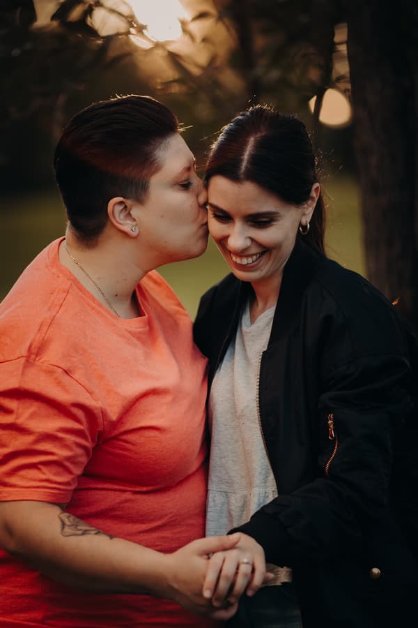 One person kissing the other's forehead while holding hands, with soft evening light.