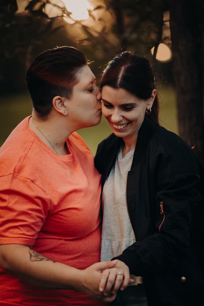 One person kissing the other's forehead while holding hands, with soft evening light.