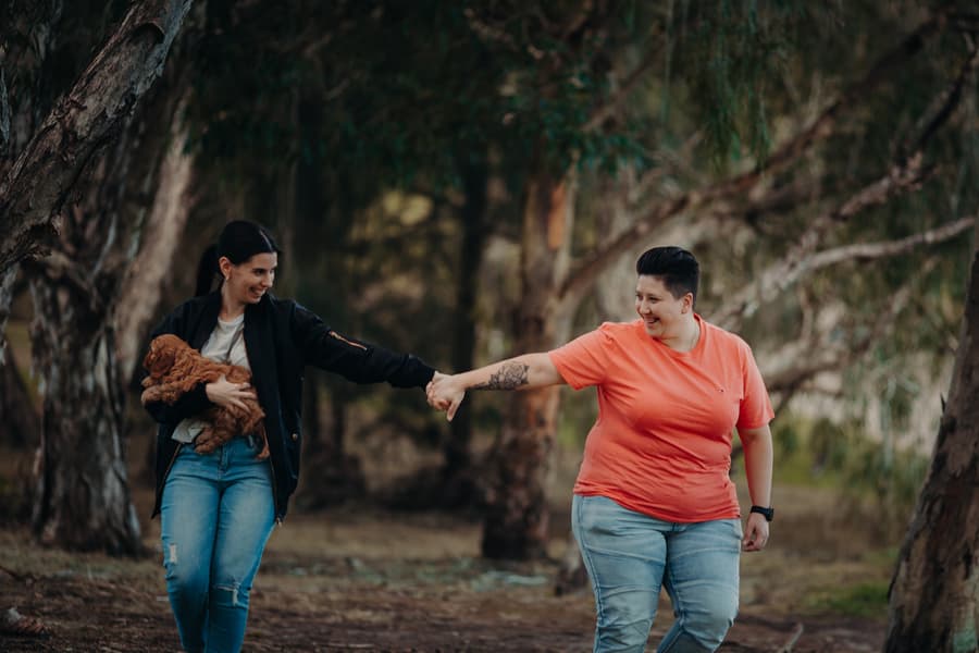 The couple walking hand in hand through a wooded area, one holding a small dog.