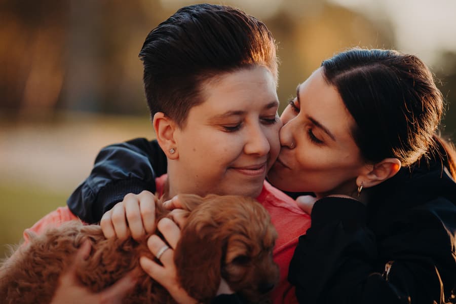 The couple embracing with a small dog, one person kissing the other's cheek.