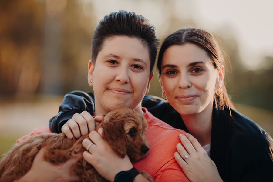 The couple smiling at the camera, holding a small dog, with soft evening light.