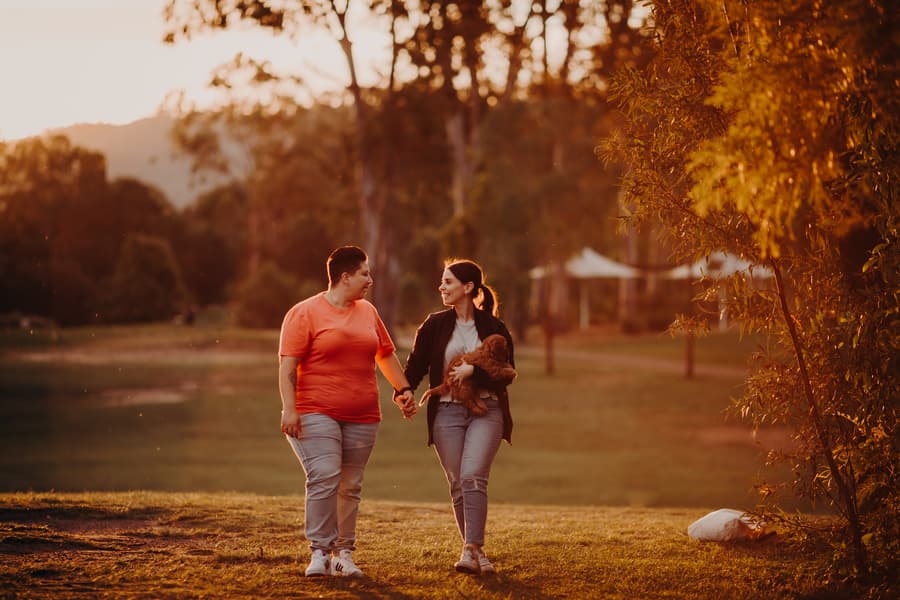 The couple walks hand in hand with a dog at Hillstone St Lucia, surrounded by trees and warm evening light.