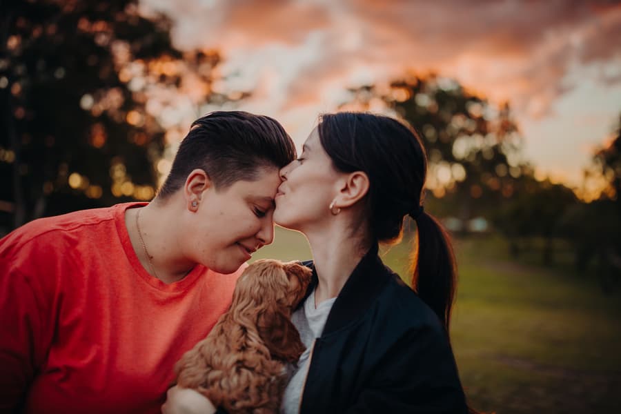 One person kisses the other's forehead while holding a dog, with a colorful sunset sky at Hillstone St Lucia.
