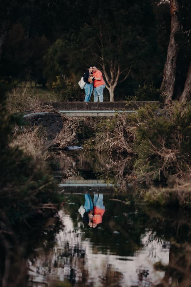 The couple stands on a small bridge, reflected in the water below, surrounded by trees at Hillstone St Lucia.