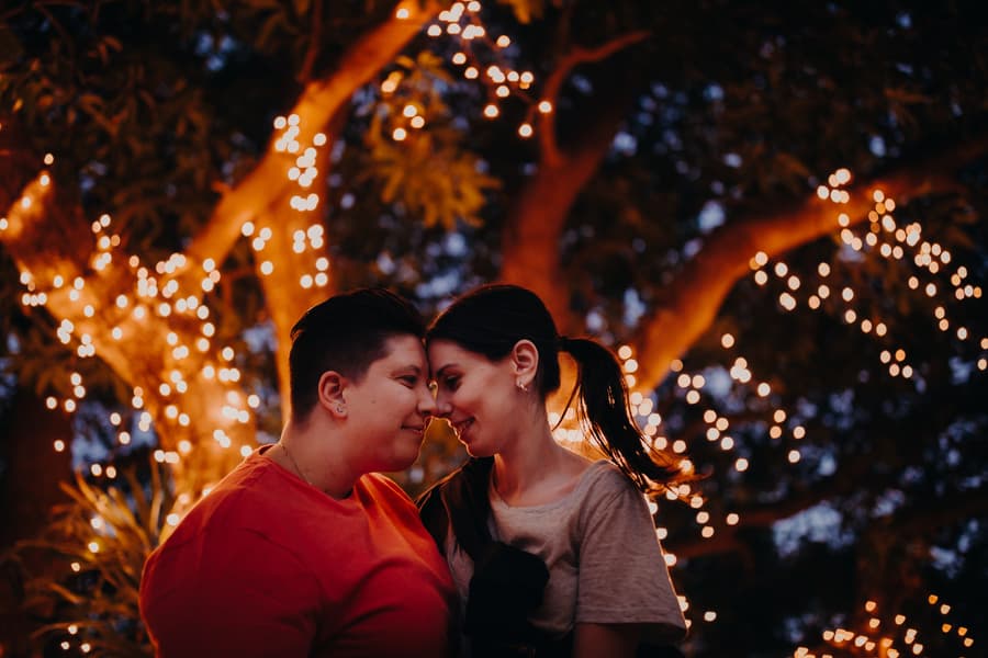 The couple stands close under a tree adorned with fairy lights, creating a romantic evening scene.