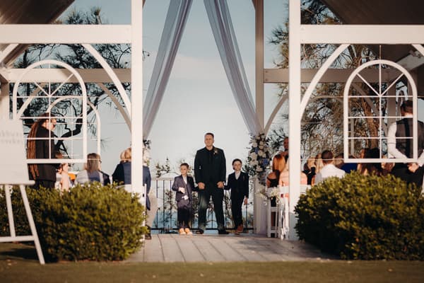 A man and two boys stand at the altar under a white pavilion structure at Sandstone Point Hotel — Pavilion, with guests seated on either side watching the ceremony.