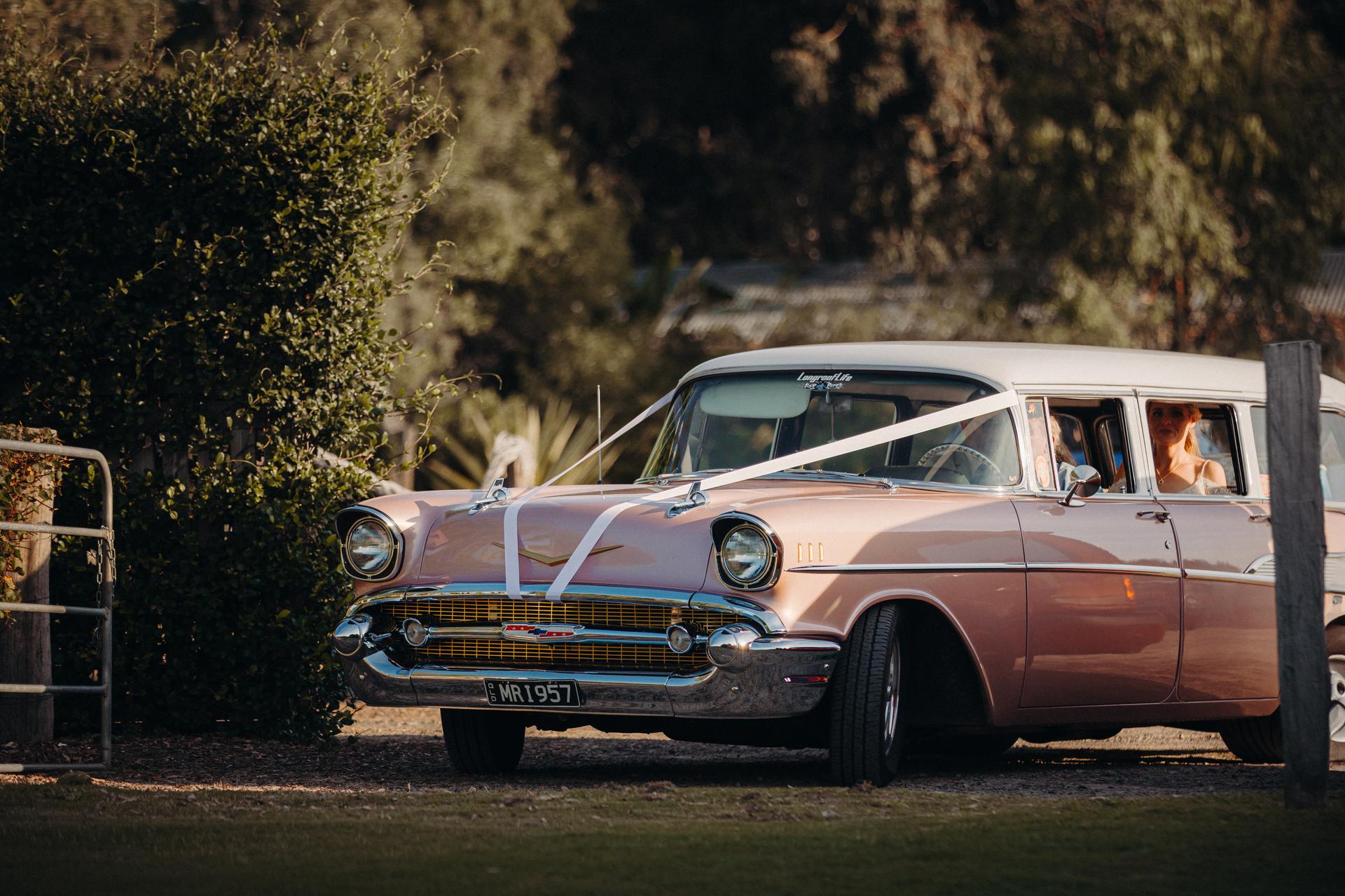A vintage pink and white car decorated with white ribbons is parked outdoors at Sandstone Point Hotel — Pavilion, with the bride visible sitting inside the car.