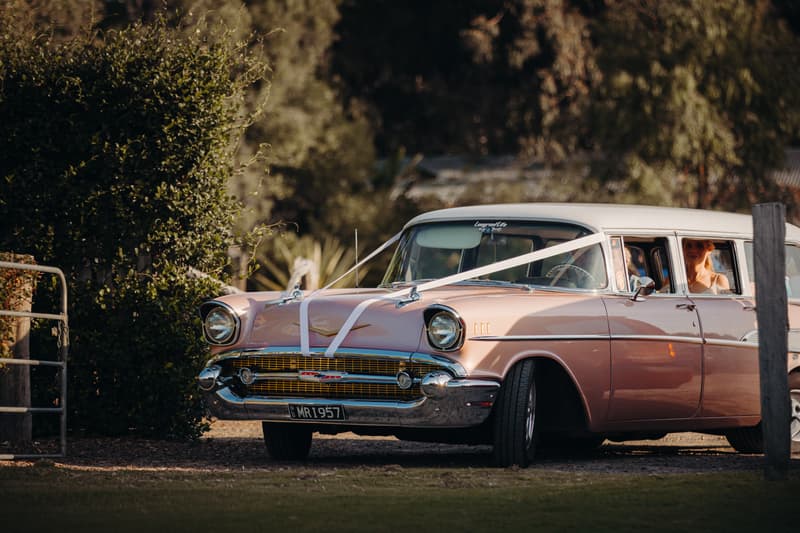 A vintage pink and white car decorated with white ribbons is parked outdoors at Sandstone Point Hotel — Pavilion, with the bride visible sitting inside the car.