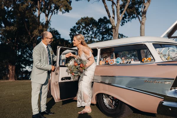 The bride Madison, holding a bouquet, steps out of a vintage car while the groom Brendon holds her hand. Elopement photography Brisbane by She Said Yes.