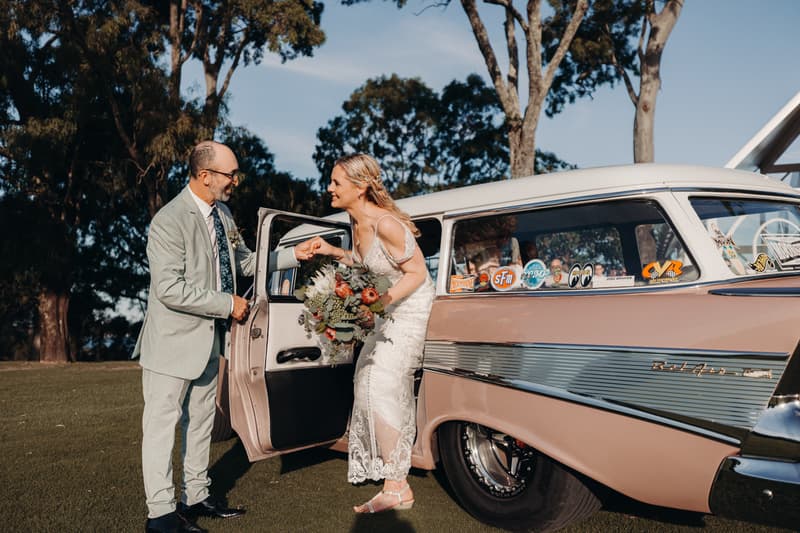 The bride Madison, holding a bouquet, steps out of a vintage car while the groom Brendon holds her hand at Sandstone Point Hotel — Pavilion.