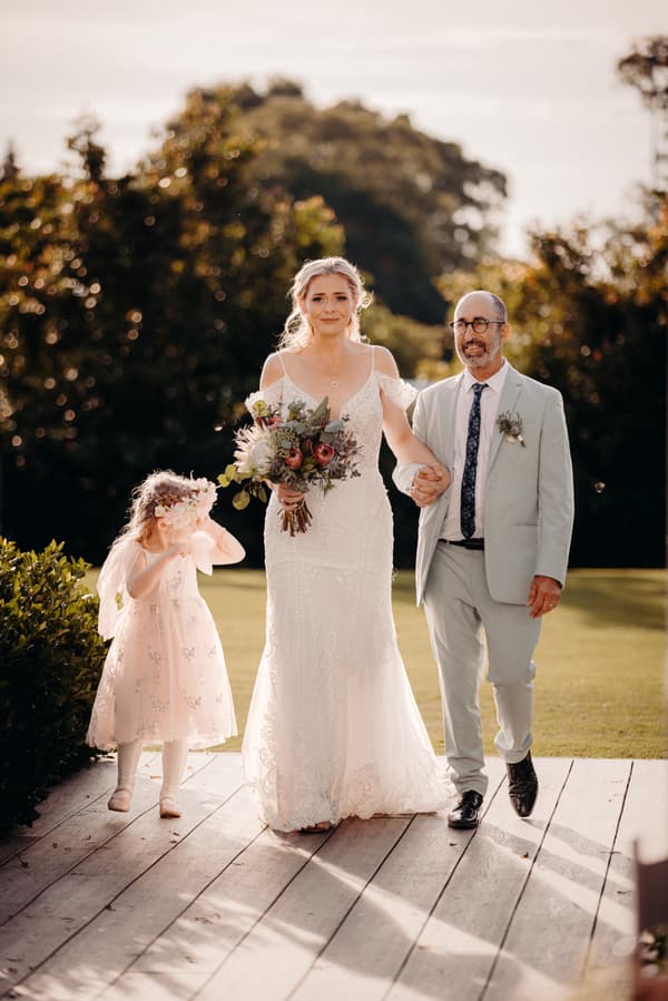 The bride Madison walks arm-in-arm with an older man, likely her father, while a flower girl in a pink dress and floral headpiece walks beside them at Sandstone Point Hotel — Pavilion.