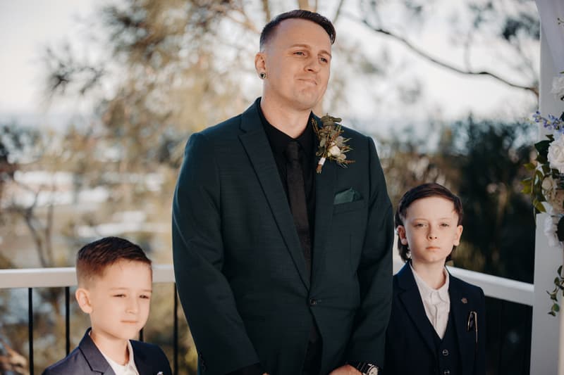 The groom stands with two young boys, all dressed in suits, at the Sandstone Point Hotel — Pavilion during the ceremony stage.