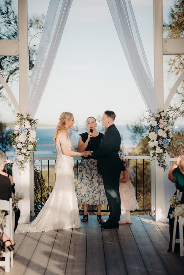 Bride Madison and groom Brendon hold hands at the ceremony stage of Sandstone Point Hotel — Pavilion, with an officiant speaking into a microphone and a flower girl standing behind them.