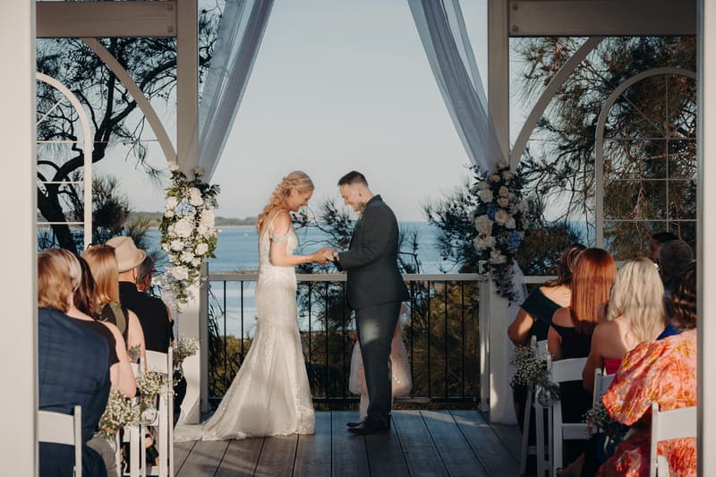 Bride Madison and groom Brendon exchange rings during their ceremony at Sandstone Point Hotel — Pavilion, with guests seated on either side and a waterfront view in the background.