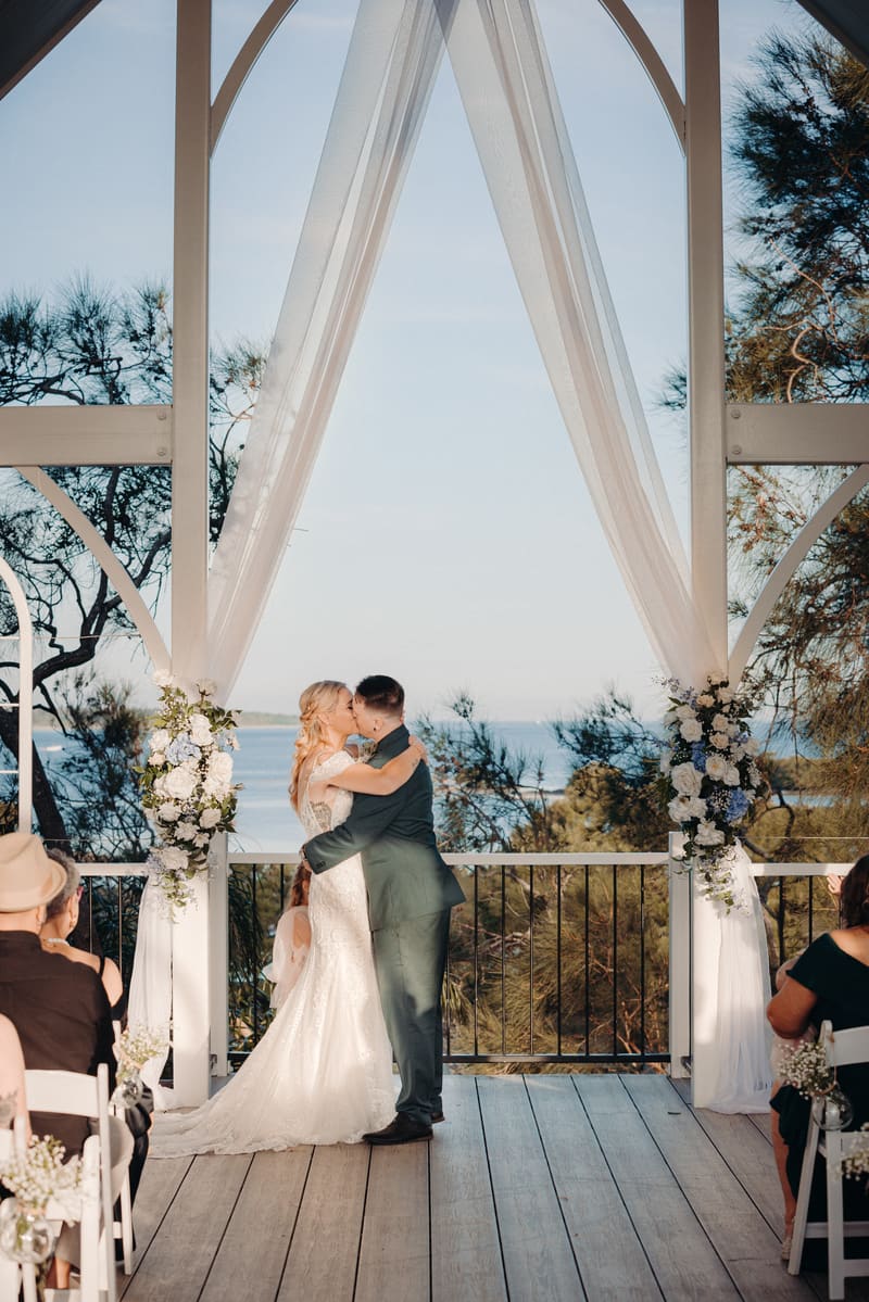 Bride Madison and groom Brendon kiss at the altar during their ceremony, framed by white drapery and floral arrangements with a waterfront view. Elopement photographer Brisbane by She Said Yes.