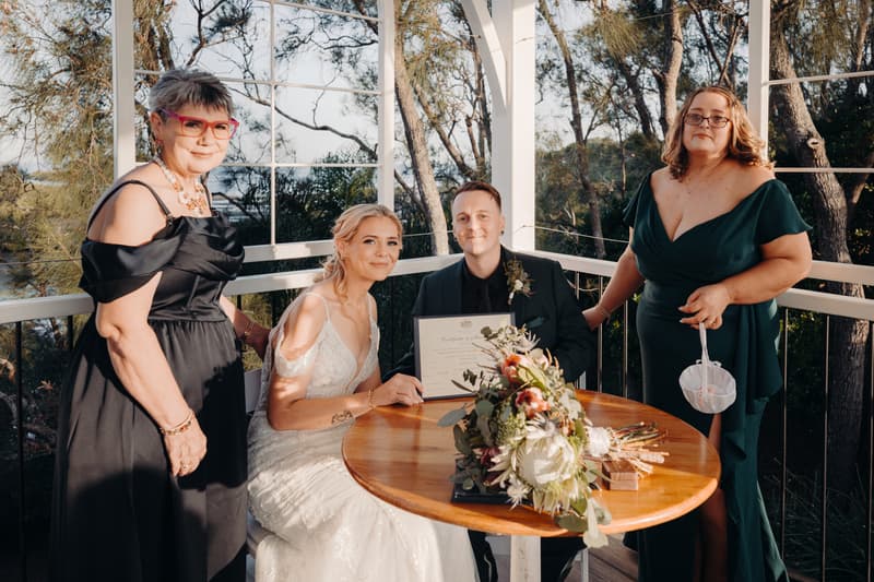 Bride Madison and groom Brendon sit at a round wooden table holding their marriage certificate at Sandstone Point Hotel — Pavilion, flanked by two women in formal dresses standing beside them.