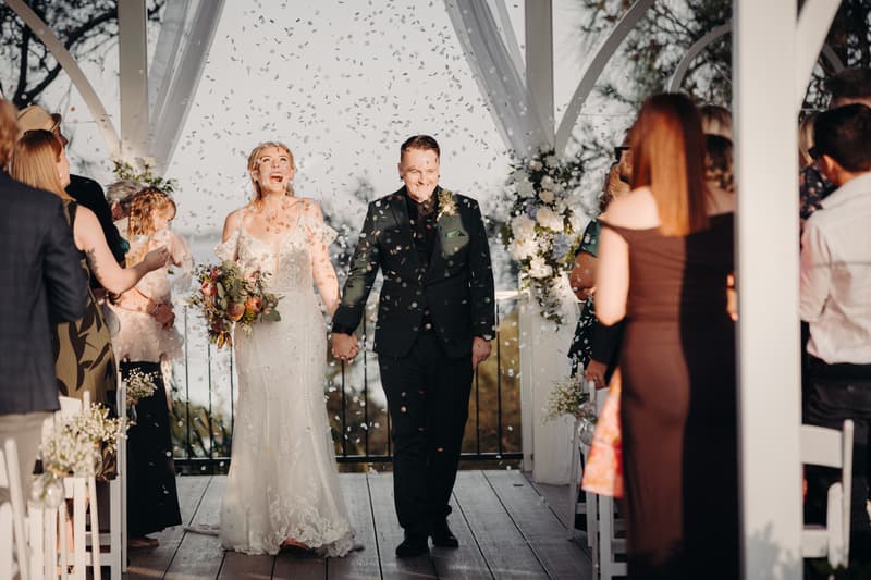 Bride Madison and groom Brendon walk down the aisle holding hands at Sandstone Point Hotel — Pavilion as guests throw confetti.