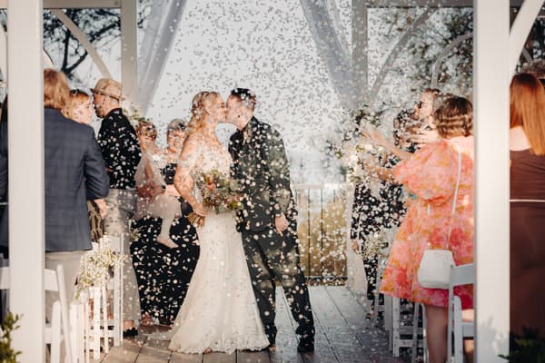 Bride Madison and groom Brendon kiss at the Sandstone Point Hotel — Pavilion ceremony stage as guests throw confetti around them.