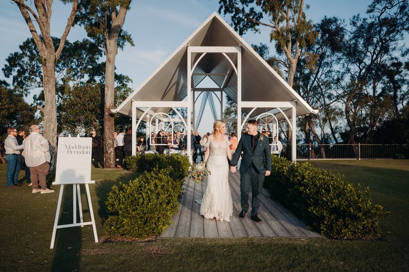 Bride Madison and groom Brendon walk hand in hand down the aisle after their ceremony at Sandstone Point Hotel — Pavilion, with guests standing and watching nearby and a wedding sign visible on the left.