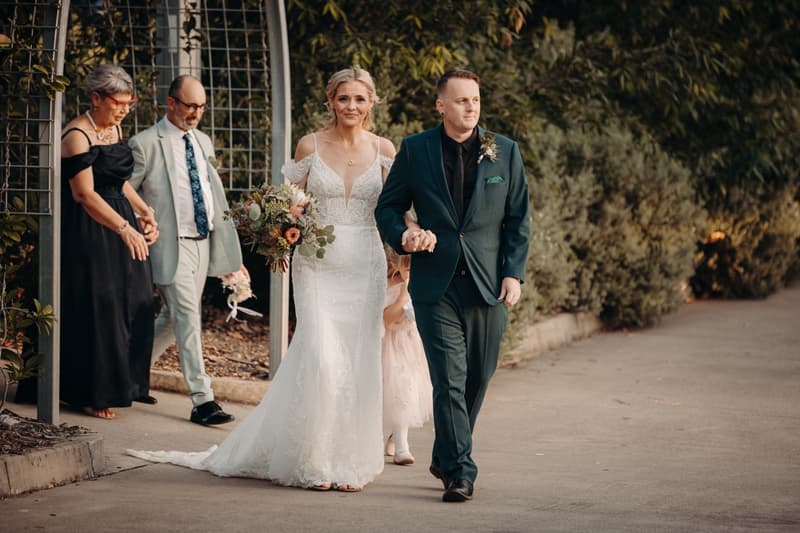 Bride Madison in a white wedding gown holding a bouquet and groom Brendon in a dark suit walk hand in hand at Sandstone Point Hotel — Pavilion, followed by two older guests and a young flower girl in a pink dress.