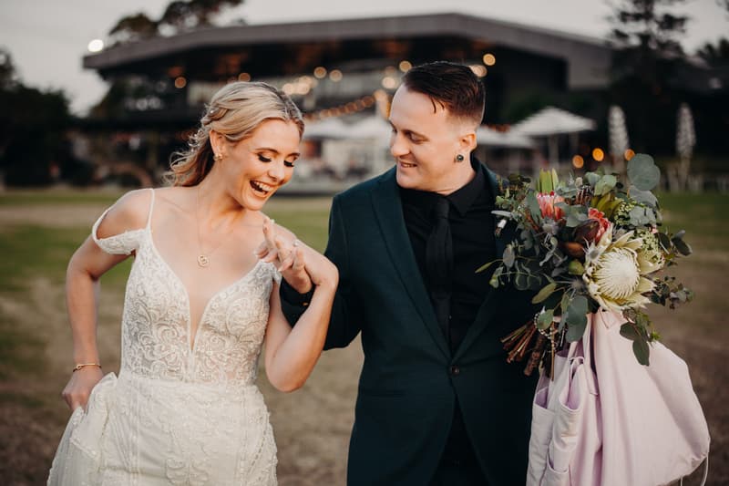 Madison the bride and Brendon the groom walk together outdoors at Sandstone Point Hotel, with Brendon holding a bouquet and a light-colored jacket while Madison smiles and looks down at their joined hands.