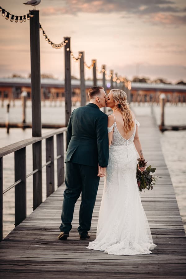 Bride Madison and groom Brendon kiss while holding hands on a wooden pier at Sandstone Point Hotel during their couple portraits session. Madison wears a white lace wedding gown and holds a bouquet, while Brendon wears a dark suit.