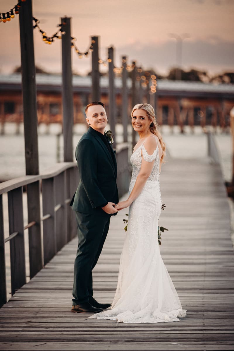 Madison and Brendon hold hands and face the camera while standing on a wooden pier. Wedding photography by She Said Yes.