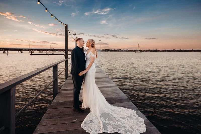 Madison and Brendon stand facing each other holding hands on a wooden pier at Sandstone Point Hotel during sunset, with string lights overhead and water surrounding the pier.