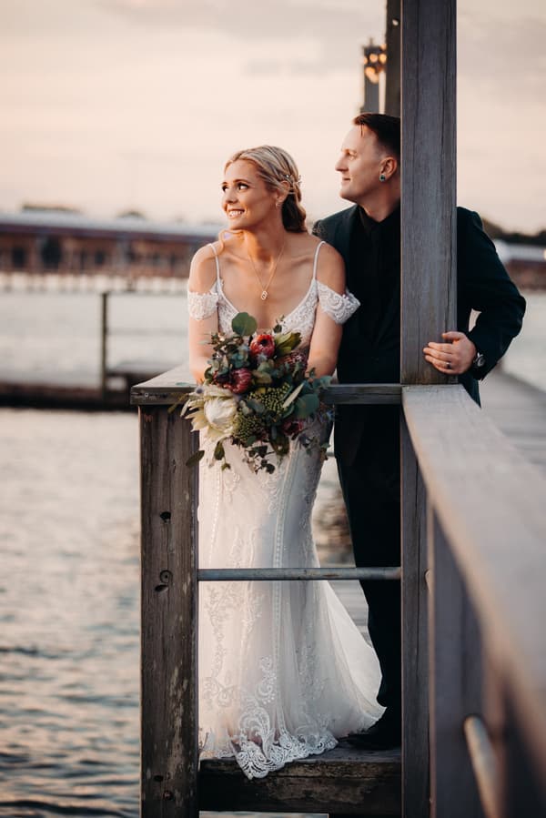Madison in a white lace wedding gown holding a bouquet and Brendon in a dark suit stand together on a wooden pier at Sandstone Point Hotel, looking towards the water during sunset.