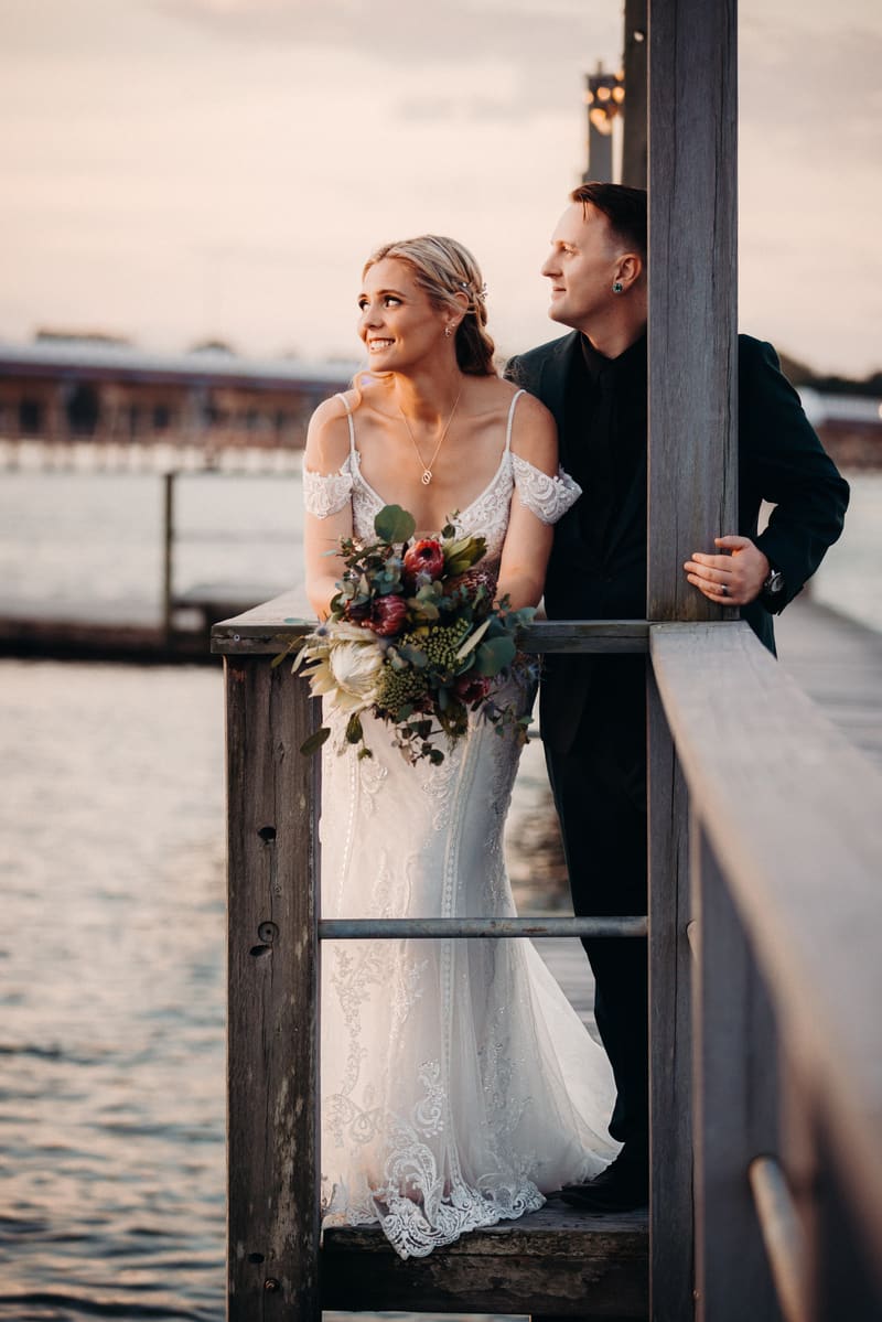 Madison in a white lace wedding gown holding a bouquet and Brendon in a dark suit stand together on a wooden pier, looking towards the water during sunset. Valentine's Day wedding photography by She Said Yes.