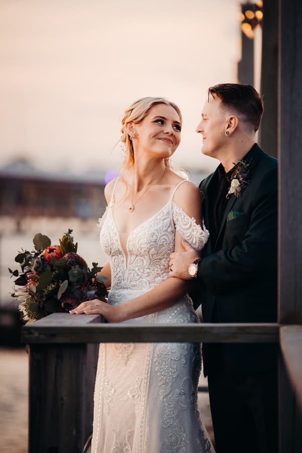 Madison and Brendon pose together during their couple portraits session at Sandstone Point Hotel, with Madison holding a bouquet and Brendon embracing her from behind.