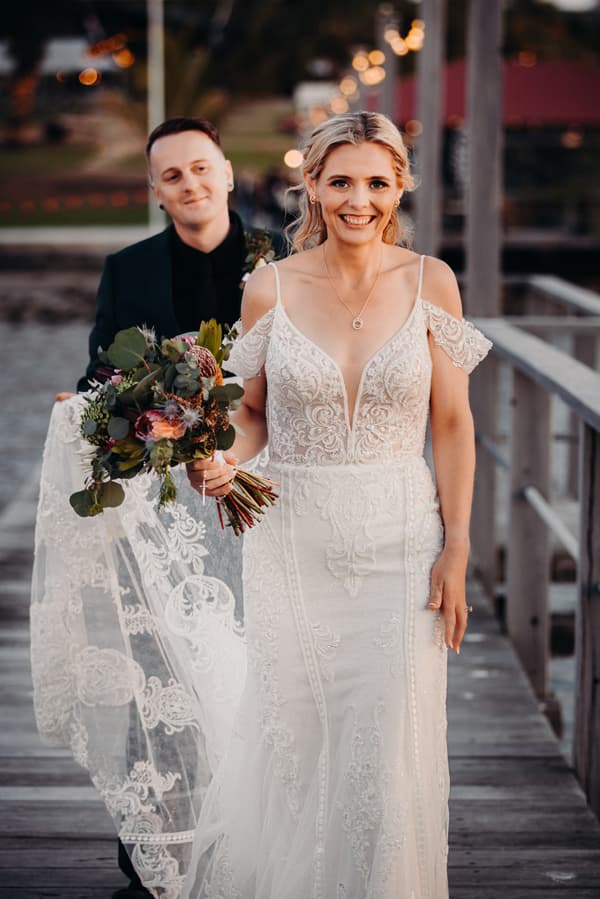 Madison in a detailed white wedding gown holding a bouquet walks on a wooden pier at Sandstone Point Hotel, with Brendon in a dark suit behind her holding her lace train.