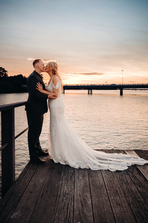 Bride Madison and groom Brendon share a kiss on a wooden pier at Sandstone Point Hotel during sunset, with water and a bridge in the background.