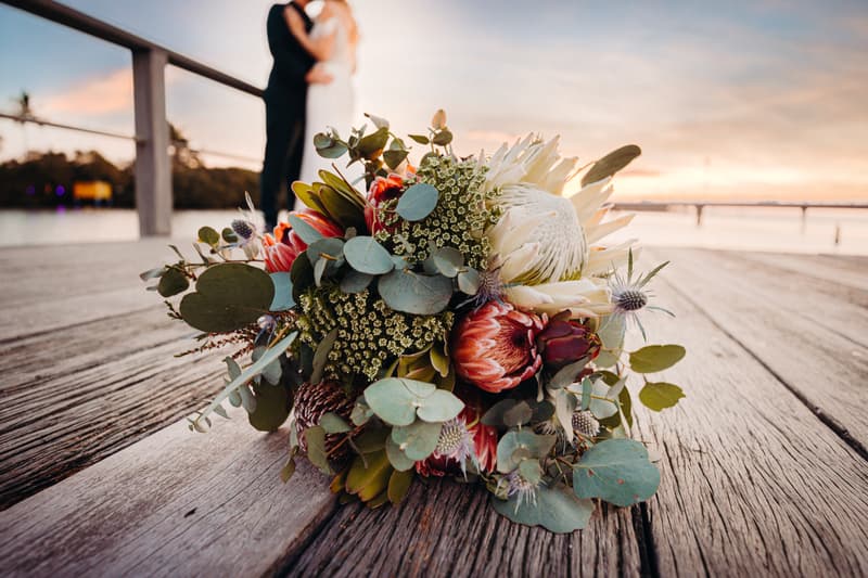 Wedding bouquet placed on wooden deck with bride and groom embracing in the background at Sandstone Point Hotel during sunset.