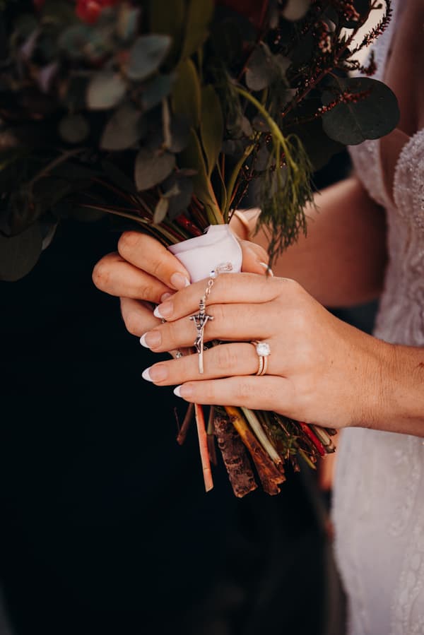 The bride Madison holds a bouquet wrapped with a white ribbon and a rosary with a crucifix, showing her wedding and engagement rings at Sandstone Point Hotel.