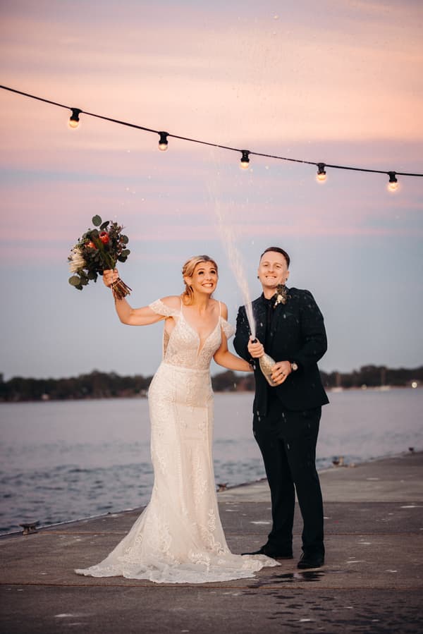Bride Madison in a white lace wedding gown holding a bouquet and groom Brendon in a black suit opening a bottle of champagne with spray at Sandstone Point Hotel waterfront during couple portraits.