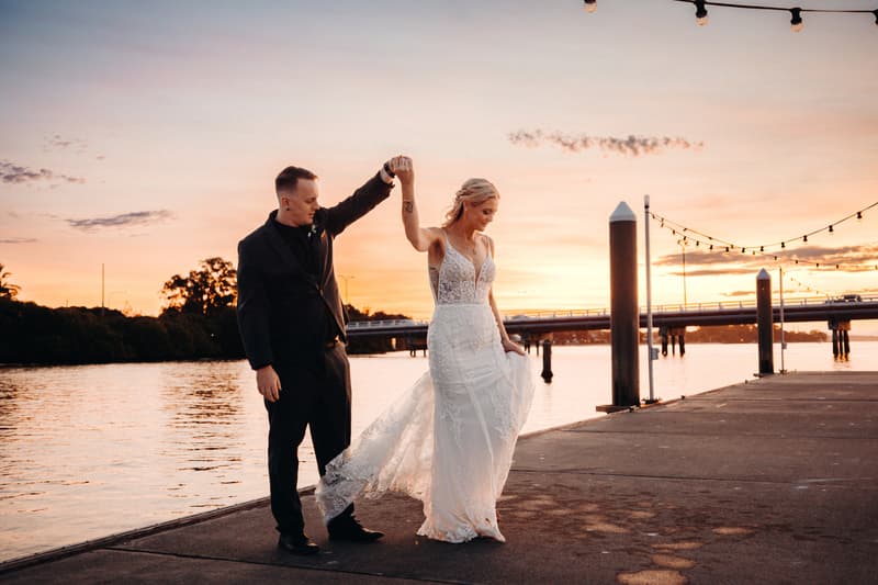 Bride Madison and groom Brendon pose together on a waterfront pier at sunset at Sandstone Point Hotel, with Brendon holding Madison's hand as she lifts her wedding dress.