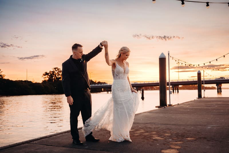 Bride Madison and groom Brendon pose together on a waterfront pier at sunset, with Brendon holding Madison's hand as she lifts her wedding dress. Award winning wedding photographer by She Said Yes.
