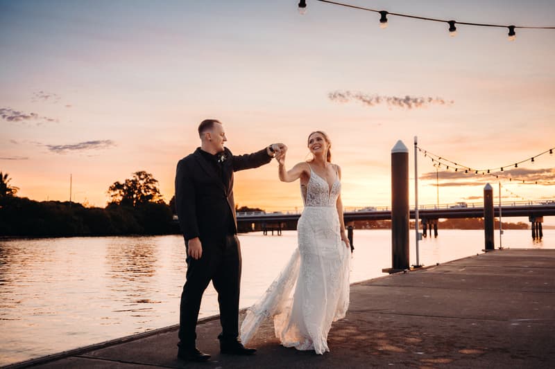 Madison in a white lace wedding gown and Brendon in a black suit hold hands and pose on a pier at sunset at Sandstone Point Hotel.