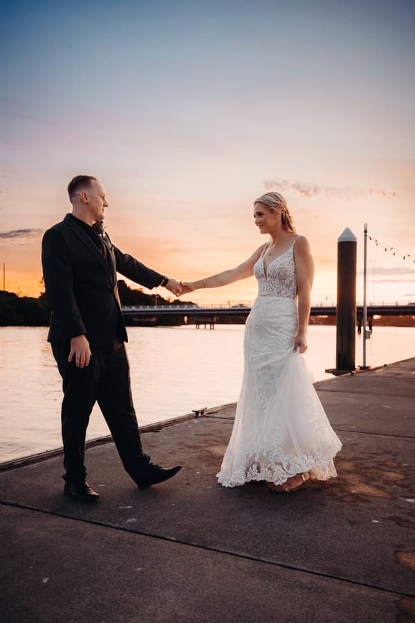 Madison in a white lace wedding dress and Brendon in a black suit hold hands on a pier at sunset at Sandstone Point Hotel.