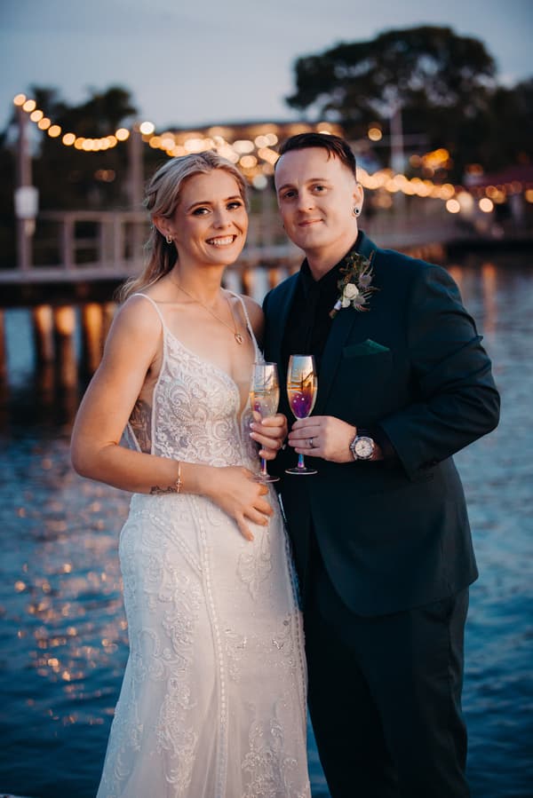 Madison in a white lace wedding gown and Brendon in a dark suit hold champagne flutes and pose together near the water at Sandstone Point Hotel during their couple portraits session.