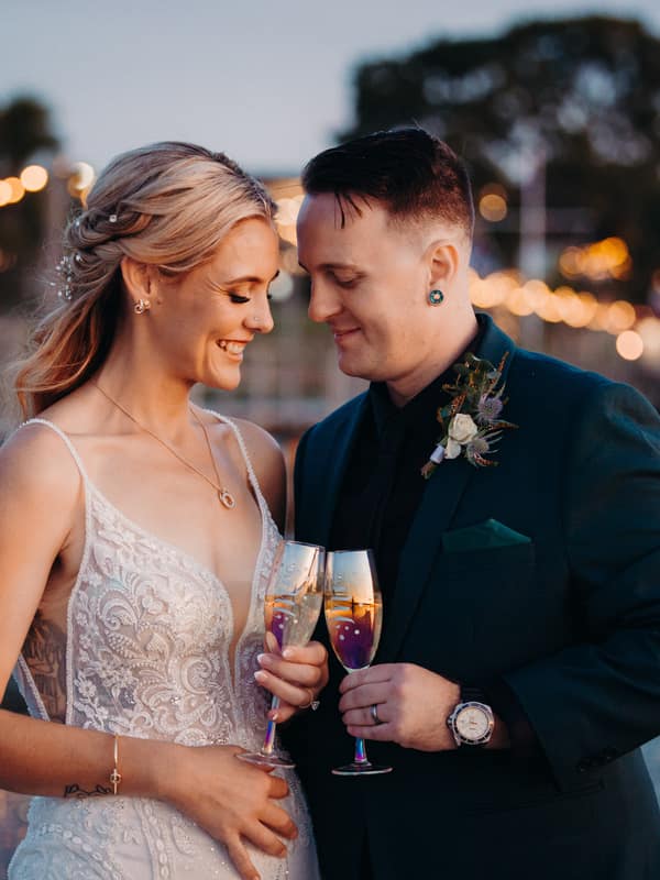 Madison and Brendon stand close together holding champagne glasses near the water at Sandstone Point Hotel during their couple portraits session.