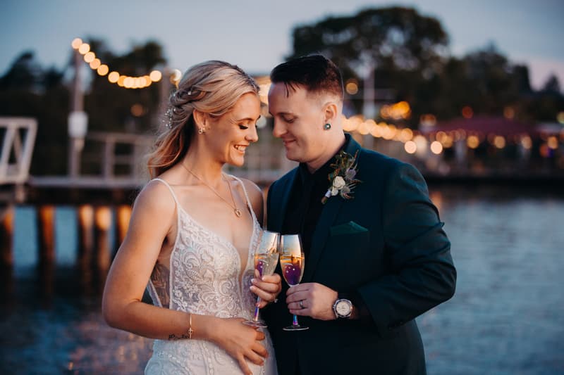 Madison and Brendon stand close together holding champagne glasses near the water at Sandstone Point Hotel during their couple portraits session.