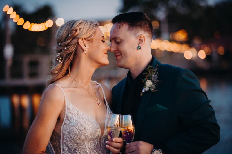 Madison the bride and Brendon the groom stand close together at Sandstone Point Hotel during their couple portraits session, holding champagne glasses with string lights blurred in the background.