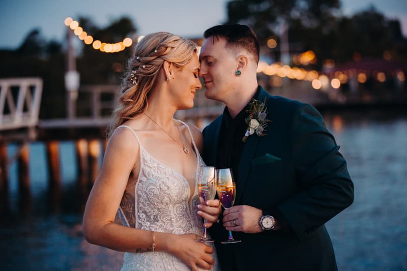 Madison and Brendon share a close moment holding champagne flutes near the water at Sandstone Point Hotel during their couple portraits session.