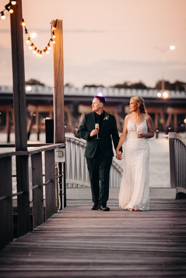 Madison and Brendon hold hands and walk along a wooden pier at Sandstone Point Hotel during their couple portraits session, each holding a glass of champagne with string lights overhead and a bridge in the background at sunset.