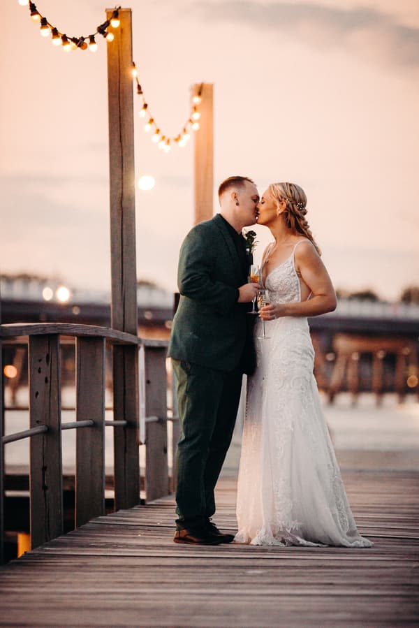 Madison and Brendon share a kiss while holding champagne glasses on a wooden pier at Sandstone Point Hotel during their couple portraits session.
