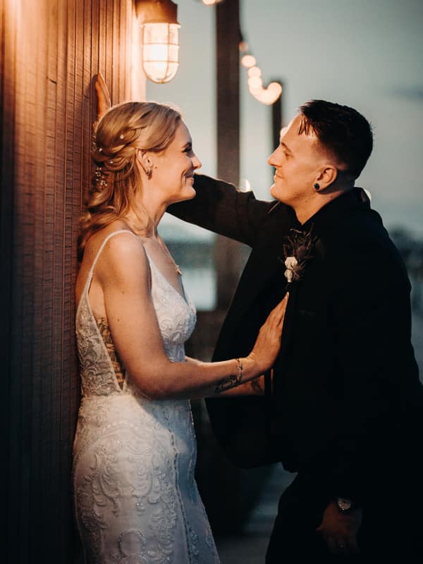 Madison in a white lace wedding dress and Brendon in a dark suit pose closely together against a wooden wall at Sandstone Point Hotel during their couple portraits session.