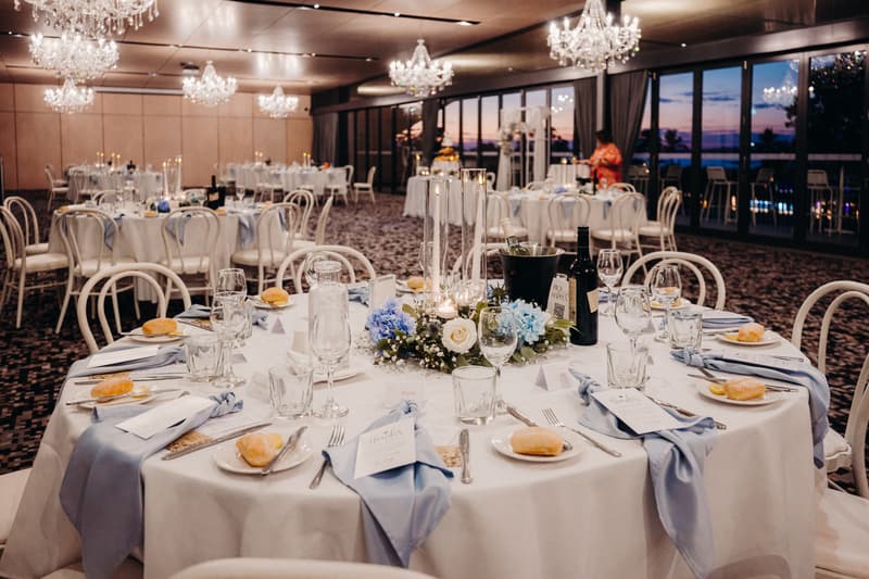 Reception tables set with white tablecloths, blue napkins, bread rolls, glassware, and floral centerpieces with candles in the Pumicestone Room at Sandstone Point Hotel during sunset.